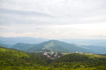 Fototapeta premium Panoramic view of rolling green mountains under a cloudy sky. highest ski resort Dragobrat of colorful roofs is nestled in a valley, surrounded by dense trees. Ukrainian Carpathians