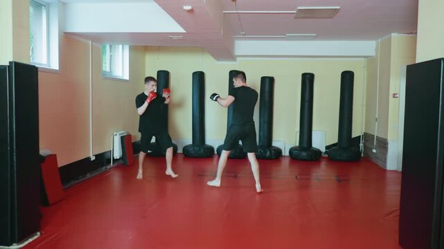 athletes sparring inside martial arts gym on red mat, practicing defensive and offensive moves in combat stance with gloves, surrounded by tall punching bags during focused training session