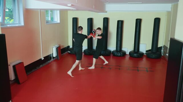 wrestlers spar barefoot on red mat in martial arts gym, one guarding face with red gloves while opponent prepares to strike, practicing combat skills surrounded by standing punching bags in training