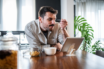 Fototapeta premium Young man eating cereal watching tablet at breakfast