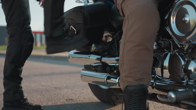 Close up of motorcycle lower section showing rider seated on chrome engine bike with tan pants and black boots while passenger begins climbing on, captured with urban background