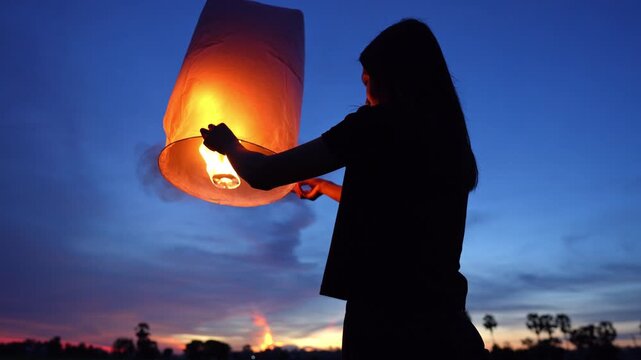 Slow motion video. Woman releases a sky lantern into the night sky, symbolizing hope, celebration and peace, during a traditional Asian festival, under a dark background light.