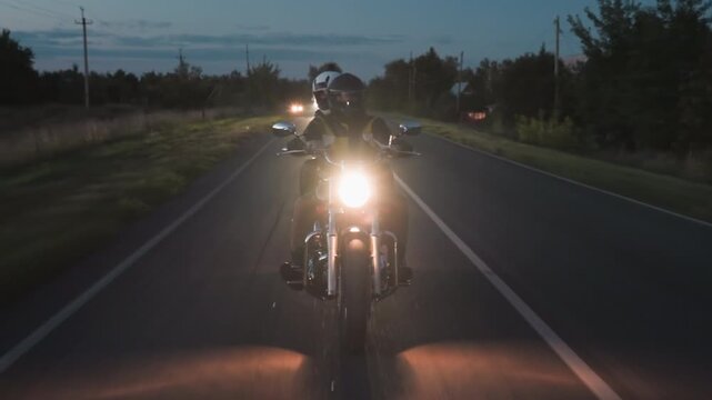 Tourist on motorcycle road trip rides through rural setting during evening, illuminated by strong headlamp, with cars following behind casting headlights across countryside road lined with trees