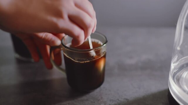 Close up of hand stirring hot coffee inside transparent glass mug on kitchen table, blending liquid with spoon, creating motion swirl while preparing fresh drink for morning refreshment