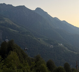Cable cars travel above lush green hills as the sun sets behind majestic mountains
