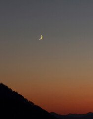 A slender crescent moon hangs in the twilight sky above dark silhouettes of mountains