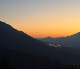 A beautiful sunset casts warm colors over the mountains as a crescent moon appears in the sky