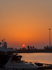 Sunset over a marina paints the sky orange and yellow, with boats and palm trees in view