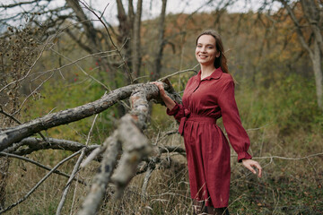 Smiling woman in a red dress leans on a fallen tree in an autumn woodland, captured with an authentic, credible feel to emphasize natural pose, textured bark and emotive connection to nature.