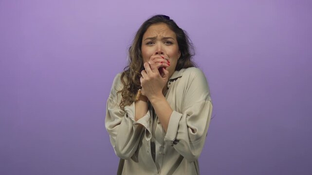 Young woman shows frightened expression and hands clasped near face in a purple studio setting; fear.
