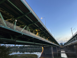 Obraz premium Warsaw, Poland - September 24, 2025: Underneath a large green bridge at dusk, showcasing its structural design and reflections on the water below, highlighting urban architecture and engineering