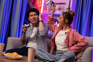 Naklejka premium Sitting sofa of happy couple playing video game with joysticks, smiling woman playing challenge, raising fist up to be winner the challenge at modern neon blub light living room at home. Infobahn.