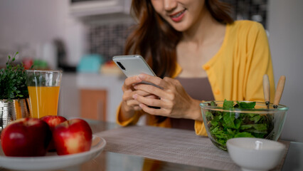 Young Asian woman enjoys digital connection at home using smartphone healthy breakfast lunch meal fresh fruit salad