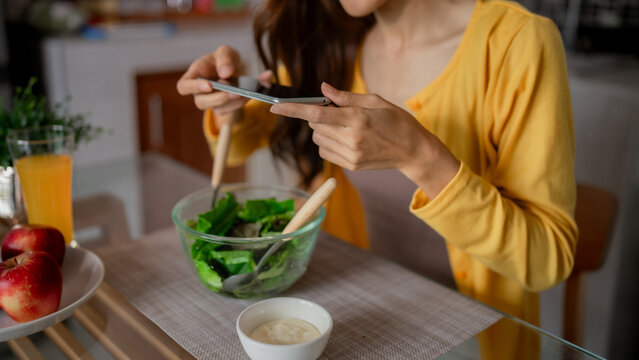 Asian adult woman photographs healthy green salad meal home kitchen promoting wellness food lifestyle content creation via smartphone