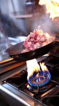 Dynamic vertical shot of a chef saut&eacute;ing meat with fire and smoke on a gas stove. Perfect for food ads, cooking shows, restaurant promos, and cinematic kitchen scenes.