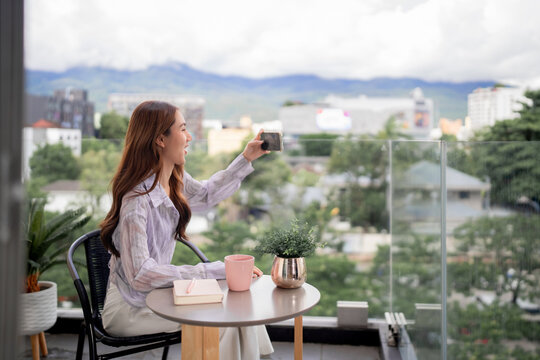Asian adult woman captures city mountain view smartphone balcony home apartment relaxed moment enjoying urban landscape outdoor living photography - Powered by Adobe