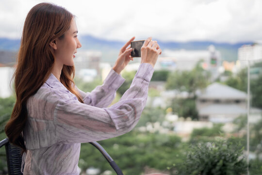 Asian adult woman captures outdoor balcony photo using smartphone enjoying scenic cityscape view creating digital memory