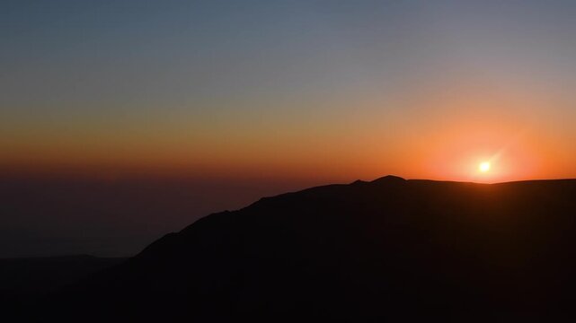 A time lapse of a vibrant sunset over the majestic Hajar Mountains in Oman. The footage captures the sun dipping below the rugged peaks, casting a warm, golden glow across the landscape as the sky tra