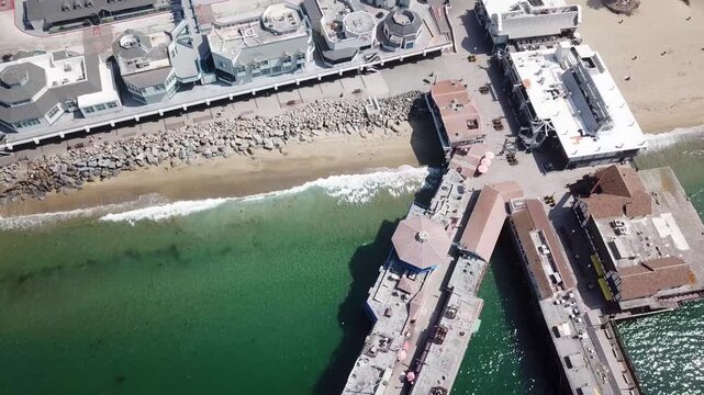 Top down drone shot tracking along the shoreline beside Redondo Beach horseshoe pier. Green clear water laps against the sandy cove and rock revetment with waterfront buildings and walkways in view