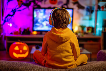 Child in yellow hoodie playing video games with headphones beside glowing pumpkin during Halloween night at cozy living room