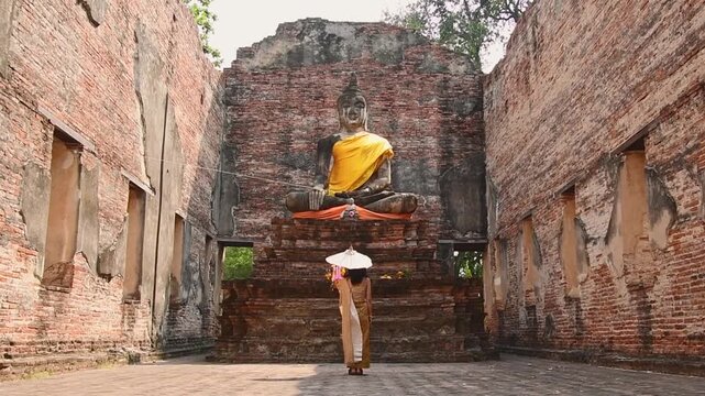 Thailand, Ayutthaya - march 15th, 2023: Woman in traditional Thai dress holding umbrella sightseeing visit ancient Buddha statue.Ayutthaya historical park. Concept heritage, spirituality, thai culture