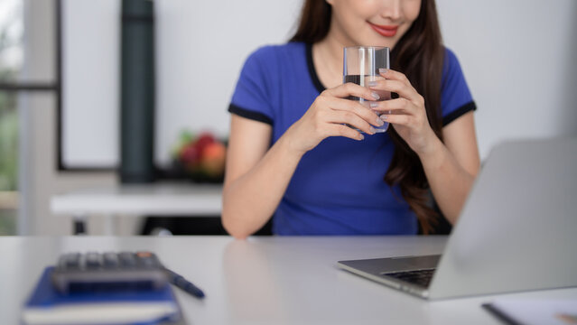 Adult Asian businesswoman drinking water for hydration and wellness at modern home office desk during work maintaining healthy lifestyle