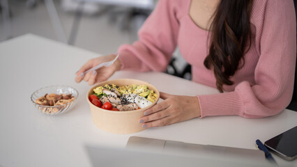 Adult Asian woman worker eating healthy salad lunch at office desk for wellness and productivity...