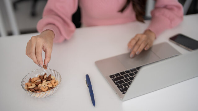 Adult Asian woman hands pick healthy snack nuts work laptop home office remote productivity well being