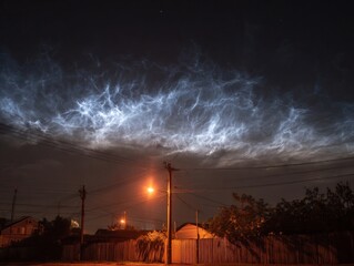 Bright noctilucent clouds illuminate the dark night sky over a suburban street with houses and power lines.