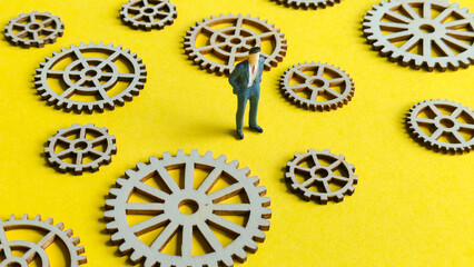Miniature businessman standing among scattered wooden gears on a bright yellow background, symbolizing complex system management, strategy, and business processes.