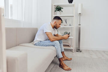 Thoughtful man sitting on a couch, deeply engaged in his smartphone while relaxing in a stylish, modern living room atmosphere full of natural light.