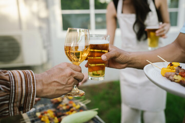 Close-up of friends clinking glasses of beer and wine during an outdoor barbecue party, celebrating and enjoying good times together.
