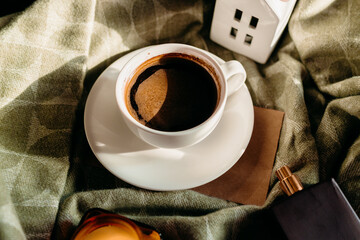 White coffee cup on green plaid blanket under sunlight. Candle adds warmth and cozy atmosphere.