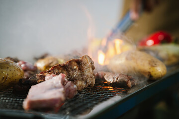 Close-up of grilled meat, potatoes, and vegetables cooking on a barbecue grill with smoke rising, showing delicious outdoor food preparation.