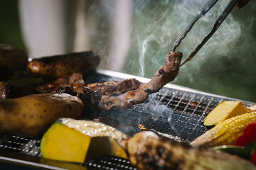 Close-up of grilled meat, potatoes, and vegetables cooking on a barbecue grill with smoke rising, showing delicious outdoor food preparation.