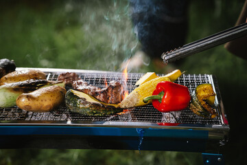 Close-up of grilled meat, potatoes, and vegetables cooking on a barbecue grill with smoke rising, showing delicious outdoor food preparation.