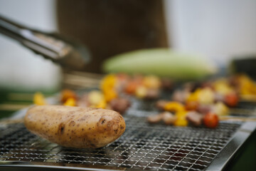 Close-up of grilled meat, potatoes, and vegetables cooking on a barbecue grill with smoke rising, showing delicious outdoor food preparation.