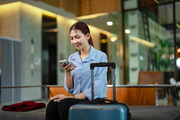 Female traveler using mobile phone for online check-in and digital payment at hotel, representing convenience, connectivity, and modern travel experience with suitcase beside her.