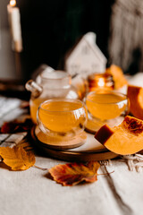 Steaming pumpkin tea in two transparent cups on wooden table with autumn leaves and warm candlelight.