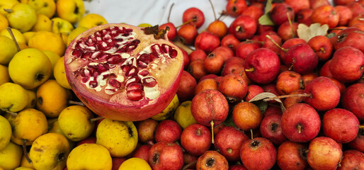 Freshly cut pomegranate with its red seeds is displayed among piles of yellow apples and small red crabapples at a Spanish market. Natural abundance.