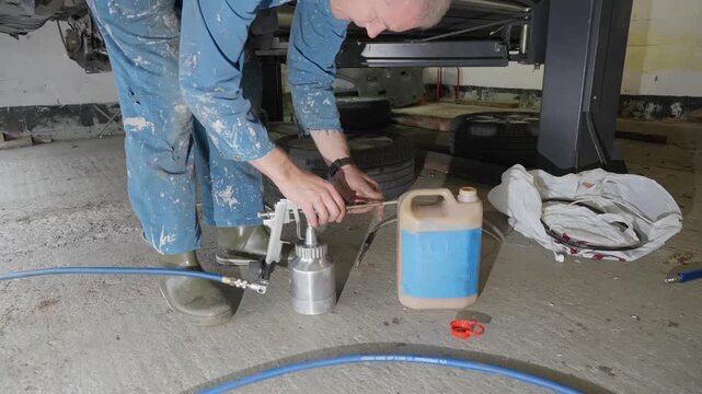 Man prepares to apply lanolin rust prevention spray to car underside, testing spray gun flow with hose and angled nozzle attached.