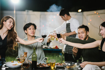 Group of friends having a fun backyard barbecue party at night, grilling food, chatting, and enjoying drinks together in a joyful atmosphere.