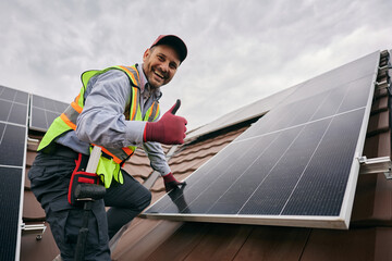 Naklejka na ściany i meble Happy worker showing thumb up while installing solar panels on roof and looking at camera