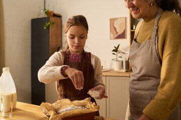 Caucasian girl sprinkling ingredients on dough while standing next to middle aged Caucasian woman in kitchen, both wearing aprons and preparing food together at counter