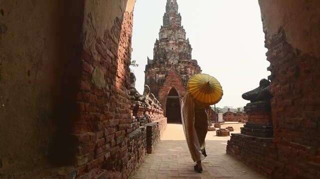 Ayutthaya, Thailand &ndash; March 2023: Woman in traditional Thai dress holding yellow umbrella walking by headless Buddha statues at Wat Chaiwatthanaram, UNESCO World Heritage Site symbol of ancient Siam.