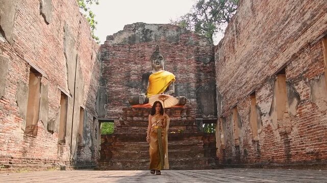 Thailand, Ayutthaya - march 15th, 2023: Woman in traditional Thai dress holding umbrella sightseeing visit ancient Buddha statue.Ayutthaya historical park. Concept heritage, spirituality, thai culture