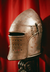 A metal helmet from medieval times rests against a rich red background. Its intricate design features ventilation holes and decorative chains.