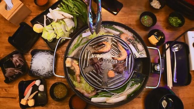 Top view of traditional Thai BBQ hotpot (Moo Krata) with mixed seafood, octopus, shrimp, fish balls, noodles, mushrooms, cabbage, corn, garlic, and dipping sauces on wooden table in Bangkok, Thailand.