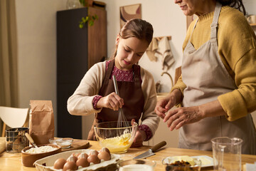 Caucasian child girl whisking batter in glass bowl while middle aged Caucasian woman assisting in kitchen, both wearing aprons, preparing ingredients for baking together