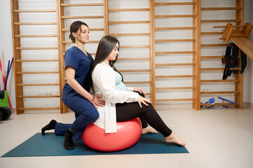 Pregnant woman doing prenatal exercises with physiotherapist using a fitness ball. Female specialist guiding pregnant client in maternity physical therapy session.
